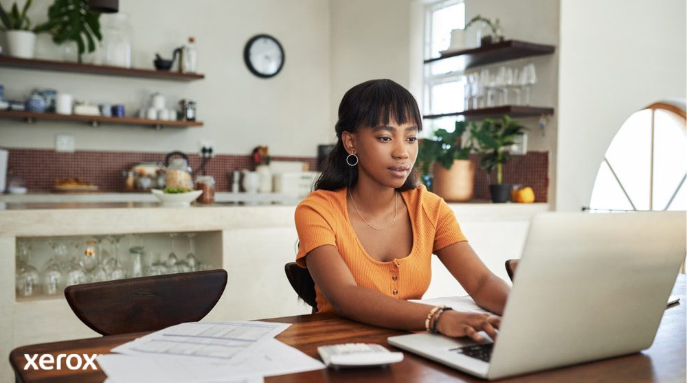 Xerox® DocuShare® Go: Ermöglichung der Remote-Arbeit Eine Person sitzt an einem Küchentisch und arbeitet konzentriert an einem Laptop. In der Nähe liegen Papiere und ein Taschenrechner. Dies veranschaulicht die Flexibilität der Heimarbeit mit effizienter Dokumentenverwaltung.