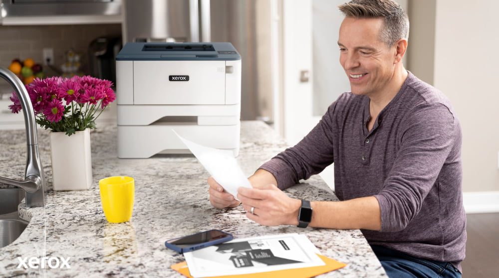 Eine Person lächelt, während sie an einer Küchentheke ein Dokument überprüft. In der Nähe stehen ein kompakter Xerox-Drucker und eine leuchtend gelbe Tasse, was auf nahtloses Drucken und Dokumentenhandling zu Hause oder in einem kleinen Büro hindeutet.