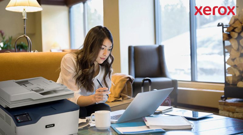Xerox® Workflow Central Platform: Überall arbeiten Eine Frau arbeitet in einem gemütlichen Büro oder einer Lounge an einem Laptop. Im Vordergrund ist ein kompakter Xerox-Drucker zu sehen, der auf eine nahtlose Integration des Druckens in digitale Arbeitsabläufe hinweist.